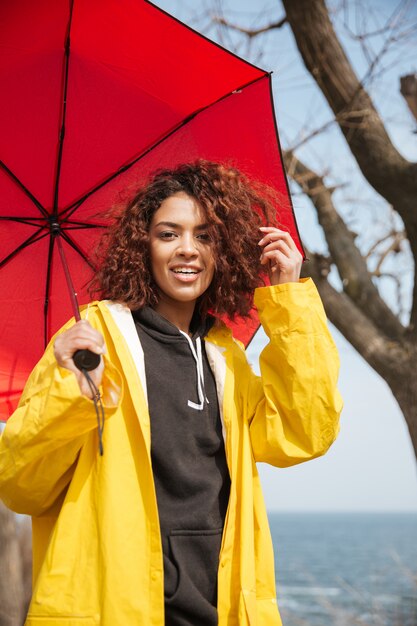 Fröhliche afrikanische lockige junge Dame, die gelben Mantel hält Regenschirm trägt