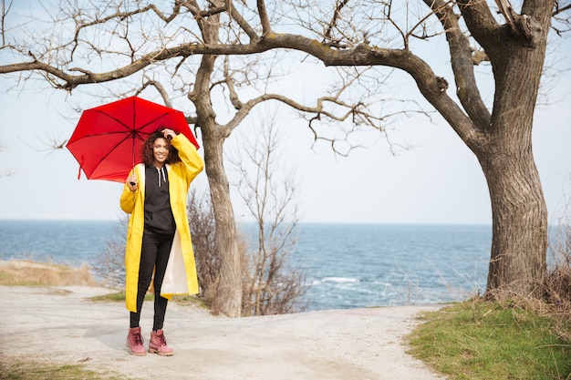 Fröhliche afrikanische lockige junge Dame, die Regenschirm hält, der draußen geht.