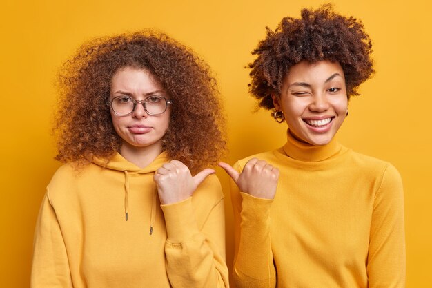 Fröhliche Afro-Amerikanerin und ihre traurige, lockige Schwester zeigen Daumen aufeinander, drücken verschiedene Emotionen aus, die beiläufig über gelber Wand isoliert gekleidet sind. Es ist sie. Zwei Frauen drinnen