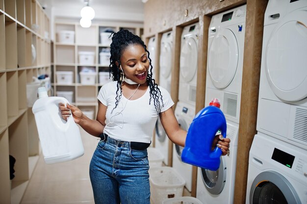 Fröhliche Afroamerikanerin hält Waschmittel in der Nähe der Waschmaschine in der Selbstbedienungswäscherei