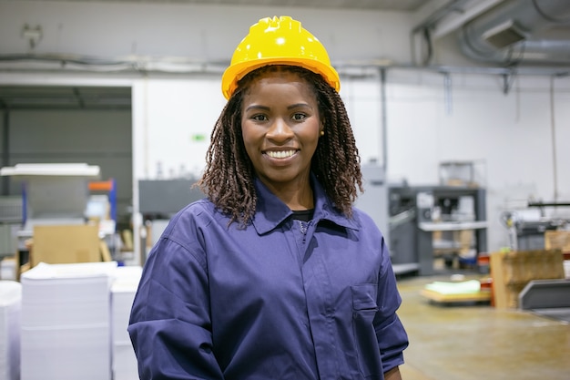 Fröhliche afroamerikanische weibliche Fabrikangestellte im Helm und insgesamt stehend auf Pflanzenboden, nach vorne schauend und lächelnd