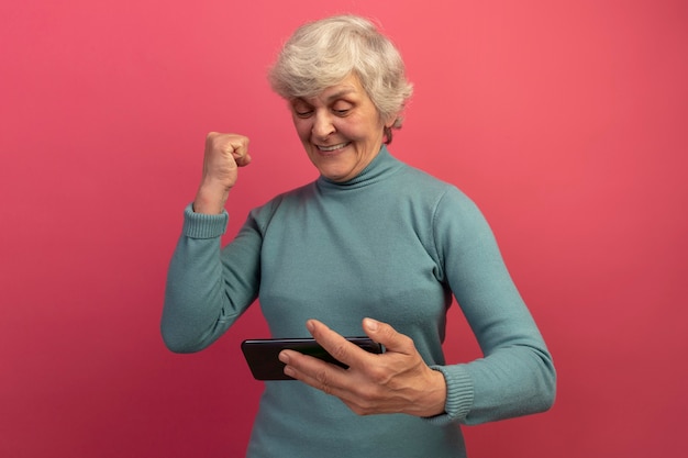 Fröhliche alte Frau mit blauem Rollkragenpullover, die das Handy hält und anschaut und dabei eine Ja-Geste macht