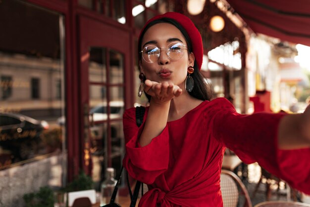 Fröhliche asiatische Dame bläst Kuss draußen Fröhliche brünette Frau in Baskenmütze und rotem Kleid macht Selfie im Straßencafé