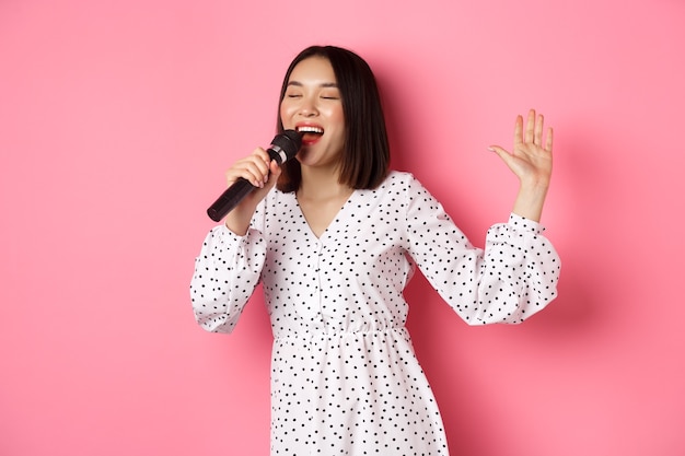 Fröhliche asiatische Frau, die im Mikrofon tanzt und singt, beim Karaoke auftritt und auf rosafarbenem Hintergrund steht. Platz kopieren