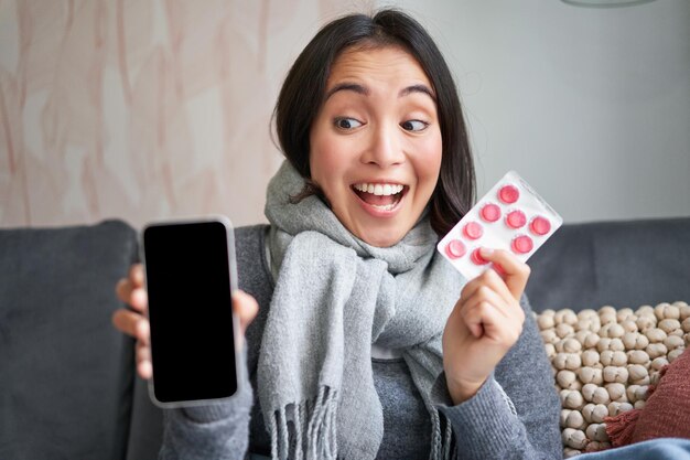 Fröhliche asiatische Frau, die sich erkältet und Smartphone-Bildschirm und Pillen zeigt, die Online-Arzt-App g empfehlen