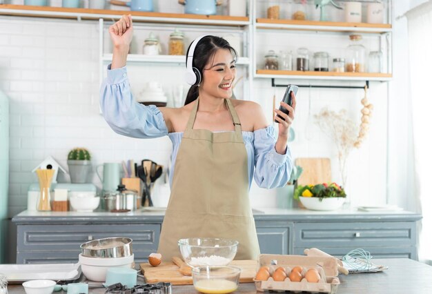Fröhliche asiatische Frau hebt Smartphone-Faust an und trägt Kopfhörer, um Musik in der Küche zu Hause zu hören