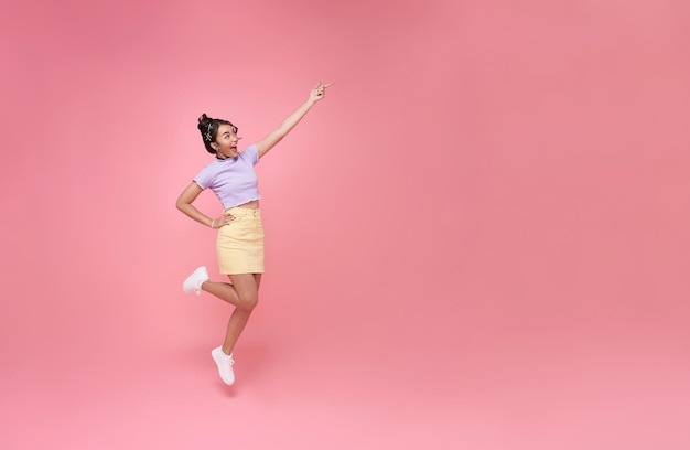 Fröhliche asiatische Frau lächelt, springt und zeigt mit dem Finger auf den Kopierraum, isoliert auf rosa Studiohintergrund