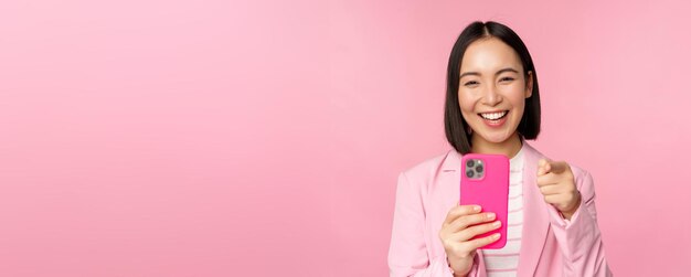 Fröhliche asiatische Geschäftsfrau lacht, zeigt mit dem Finger auf Sie und nimmt Videos auf, die Fotos auf dem Smartphone mit rosa Hintergrund des Mobiltelefons machen