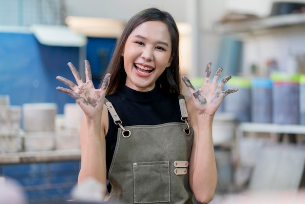 Fröhliche asiatische Keramikerin zeigt schmutzige Hand, während sie Tonvase in der Heimstudiofabrik modelliert, mit glücklich lächelndem Blick auf cameraasia weibliche Kleidungsschürze Handgestenlächeln in der Fabrik