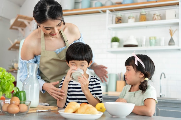Fröhliche asiatische Mutter und Sohn mit Tochter, die morgens in der Küche Milchgläser hält Genießen Sie Familienaktivitäten zusammen
