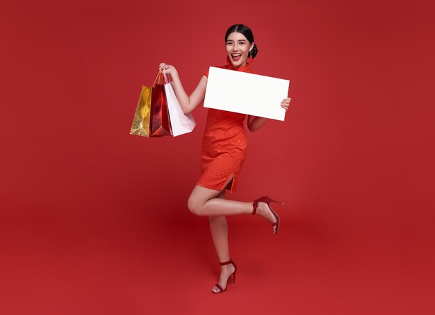 Fröhliche asiatische Shopaholic-Frau in traditionellem Cheongsam-Qipao-Kleid mit Einkaufstasche und leerem Banner in den Händen auf rotem Hintergrund.