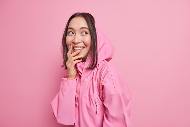 Fröhliche asiatische Teenagerin blickt Tagträume beiseite und erinnert sich an einen schönen Moment, in dem das Lächeln breit im Hoodie gekleidet ist und positive, ehrliche Emotionen ausdrückt, die über einer rosa Wand isoliert sind. Jugendlebensstil