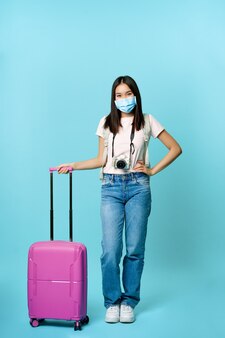 Fröhliche asiatische touristin, die in medizinischer gesichtsmaske mit koffer und kamera steht, auf tour geht, urlaub im ausland während der covid-19-pandemie, blauer hintergrund