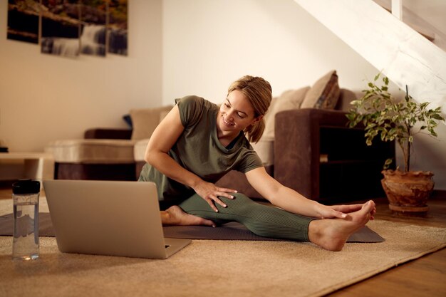 Fröhliche Athletin mit Laptop, während sie in ihrem Wohnzimmer Dehnübungen macht