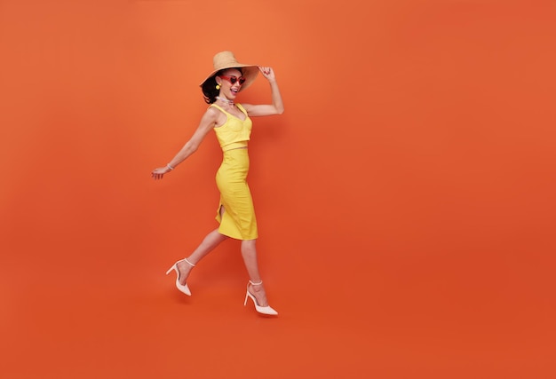Fröhliche attraktive Frau im Sommerkleid und Hut, die zu Fuß isoliert auf orangefarbenem Hintergrund des Kopierraums gehen