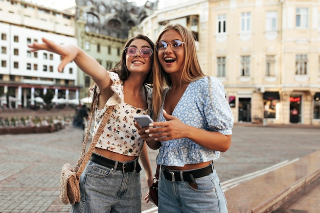 Fröhliche attraktive Frauen in stylischen Sommeroutfits blicken überrascht in die Ferne Aufgeregte blonde und brünette Mädchen in bunten Sonnenbrillen posieren im Freien und lächeln