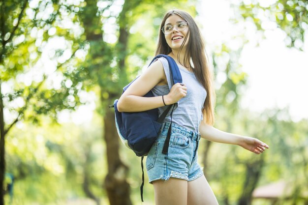 Fröhliche attraktive junge Frau mit Rucksack und Notizbüchern, die im Park stehen und lächeln