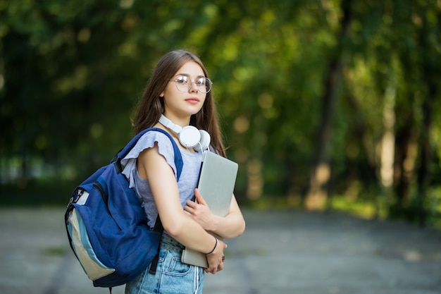 Fröhliche attraktive junge Frau mit Rucksack und Notizbüchern, die im Park stehen und lächeln