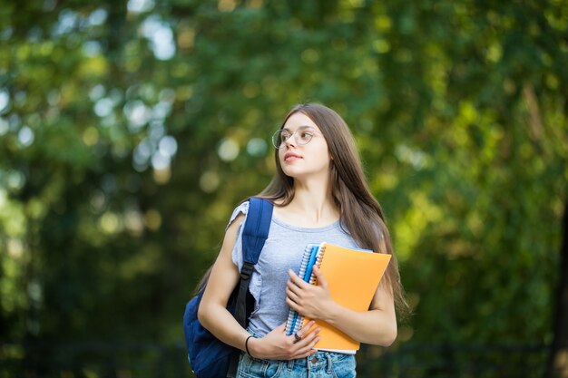 Fröhliche attraktive junge Frau mit Rucksack und Notizbüchern, die im Park stehen und lächeln