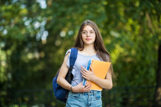 Fröhliche attraktive junge Frau mit Rucksack und Notizbüchern, die im Park stehen und lächeln