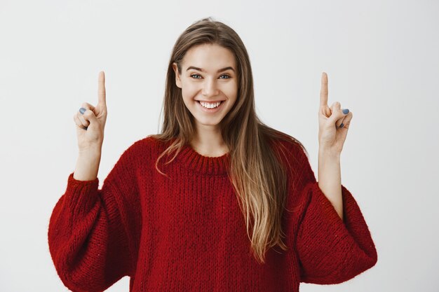 Fröhliche aufgeregte europäische Freundin, die durch Werbung großartigen Kopierraum mit uns teilt. Begeisterte glückliche Frau im stilvollen roten losen Pullover, die Finger hebend und mit breitem Lächeln nach oben zeigend