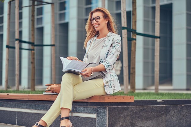 Fröhliche blonde Frau in moderner Kleidung, die mit einem Buch studiert, auf einer Bank im Park gegen einen Wolkenkratzer sitzt.