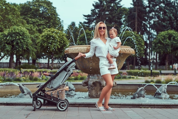 Fröhliche blonde Frau in weißer Bluse, Shorts und Sonnenbrille hält ihre kleine Tochter an den Händen und posiert in der Nähe eines Brunnens im Park.