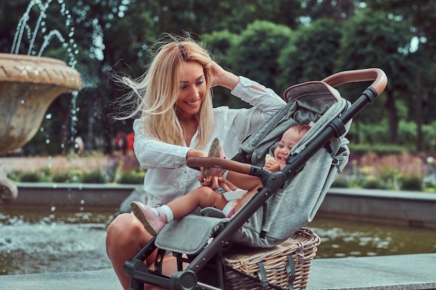 Fröhliche blonde Frau in weißer Bluse und Shorts posiert mit ihrer kleinen Tochter in einem Kinderwagen in der Nähe eines Brunnens im Stadtpark.