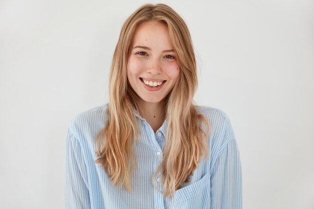 Fröhliche blonde junge Frau mit attraktivem Blick, breitem strahlendem Lächeln, gekleidet in modischem Hemd