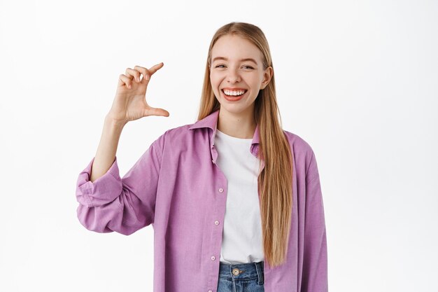 Fröhliche blonde Studentin, die kleine Größe, kleine Handgeste und ein fröhliches Lächeln zeigt, in Freizeitkleidung gegen weiße Wand stehend