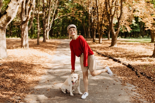 Fröhliche Blondine in stilvollem rotem Pullover und beigen Shorts, die Spaß im schönen Herbstpark mit ihrem Hund haben.