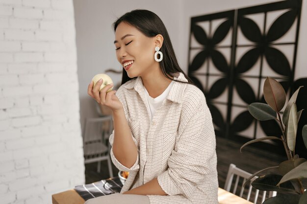 Fröhliche brünette asiatische Dame in beiger Strickjacke und Hose lächelt aufrichtig und schaut auf frischen Apfel Coole gebräunte Frau sitzt auf einem Holztisch in der Küche und hält Obst