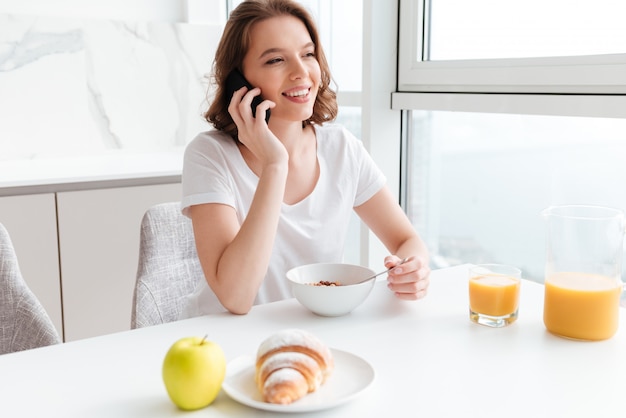 Fröhliche brünette Frau im weißen T-Shirt, das auf Handy spricht, während gesundes brekfast in der weißen Küche hat