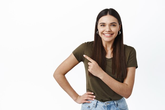 Fröhliche brünette Frau lächelt und zeigt mit dem Finger nach links auf das Promo-Banner, das Werbung in legerem Outfit zeigt, das über weißem Hintergrund steht