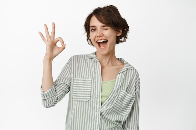 Fröhliche brünette Frau zwinkert und lächelt, zeigt ein OK-Zeichen, genehmigt etw, lobt und beglückwünscht Ihre Wahl und steht auf weißem Hintergrund