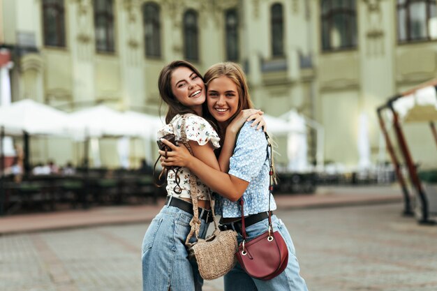 Fröhliche Brünette und junge blonde Frauen in stylischen Jeanshosen und bunten Blusen freuen sich, haben Spaß, lächeln im Freien weit