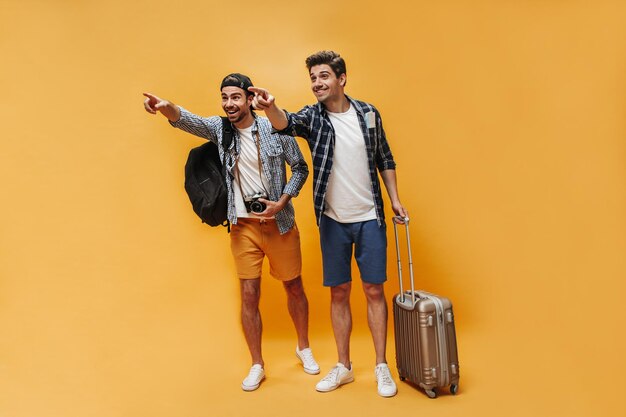 Kostenloses Foto fröhliche brunet-männer zeigen in die ferne und freuen sich reisende in karierten hemden posieren mit koffer, retro-kamera und rucksack auf orangefarbenem hintergrund