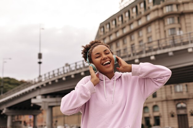 Fröhliche, coole, lockige brünette Frau in stylischem rosa Hoodie singt, lächelt und hört Musik über Kopfhörer im Freien