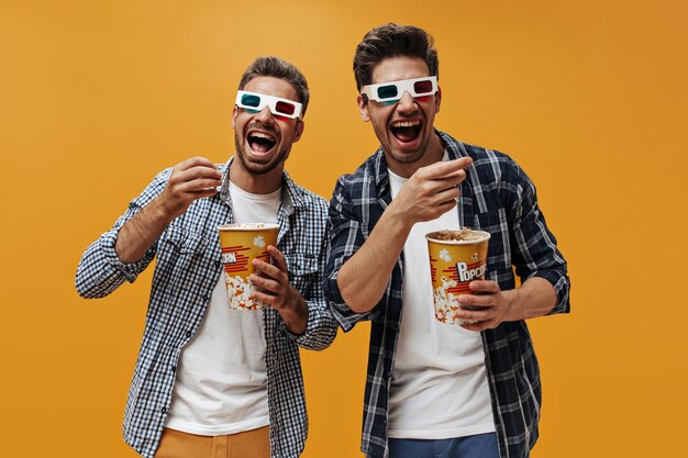 Fröhliche coole Männer in blau karierten Hemden und weißen T-Shirts sehen sich Filme in 3D-Brillen an, essen Popcorn und lachen auf orangefarbenem Hintergrund