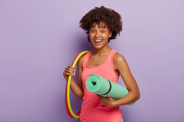 fröhliche culy haired sportliche Frau hält aufgerollte Matte, zwei Hula Hoops, trägt rosa Weste, hat Training im Fitnessstudio