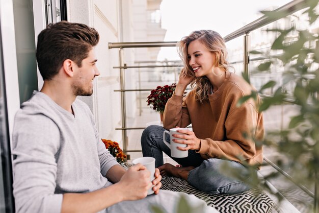 Fröhliche Dame, die mit Ehemann am Balkon spricht. Charmante junge Frau, die Tee an der Terrasse trinkt.