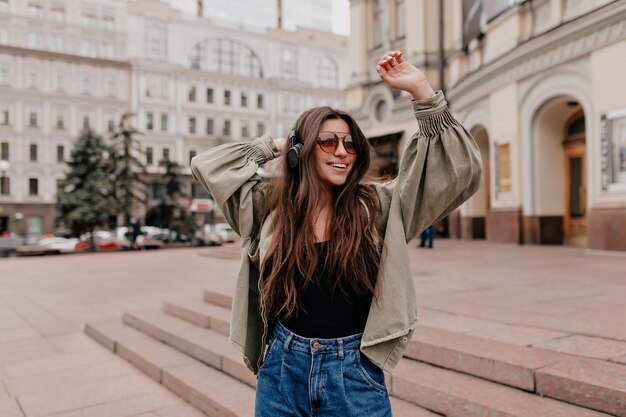 Fröhliche Dame hört Musik und tanzt in der Stadt Hübsches junges Mädchen mit langen dunklen Haaren in Jacke und Jeans mit nacktem Make-up, das lächelt und die Augen verengt, während es den Kopf winkt