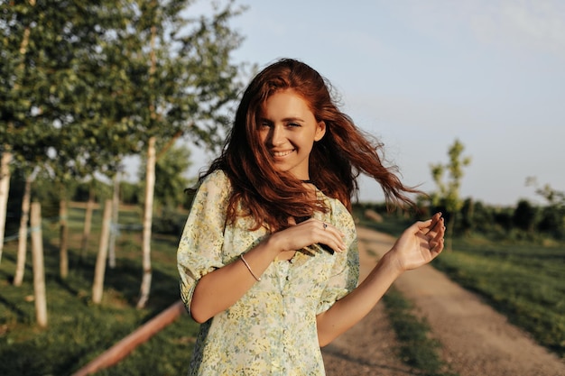 Fröhliche Dame mit langer roter Frisur und Verband am Hals im modischen Sommerkleid lächelt und schaut in die Kamera im Freien