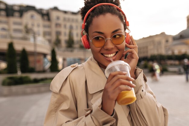 Fröhliche dunkelhäutige Frau in beigem Trenchcoat und Brille trinkt Orangensaft, hört Musik über Kopfhörer und lächelt draußen