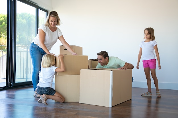 Fröhliche Eltern und zwei Mädchen, die Spaß beim Öffnen von Kisten und Auspacken in ihrer neuen leeren Wohnung haben