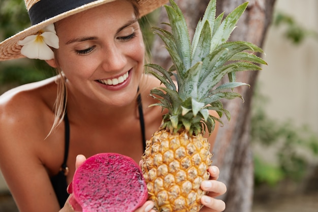 Fröhliche entzückende frau im strohhut genießt sommerferien am tropischen strand, hält exotische ananas und drachenfrucht