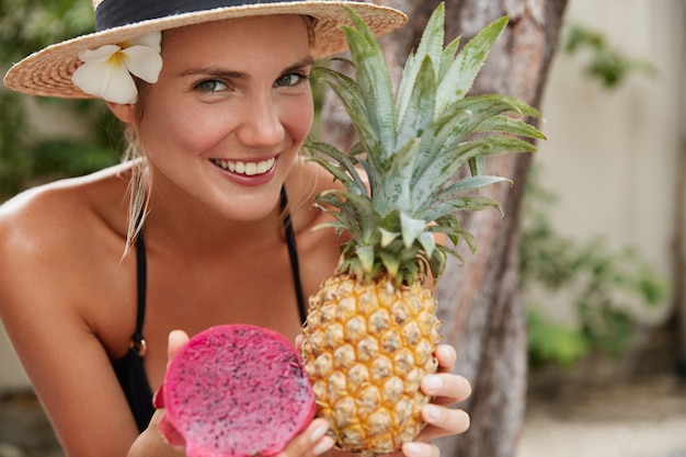 Kostenloses Foto fröhliche entzückende frau im strohhut genießt sommerferien am tropischen strand, hält exotische ananas und drachenfrucht