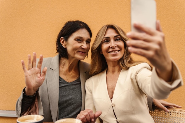 Fröhliche erwachsene kaukasische Damen machen ein Foto auf einem modernen Smartphone, während sie mit einer Tasse Kaffee auf dem Hintergrund der Wand am Tisch sitzen