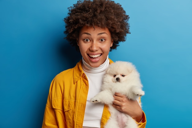 Fröhliche ethnische Frau mit Afro-Frisur, die froh ist, das Leben eines obdachlosen Welpen zu retten, hält weißen Spitz eng an sich und ist auf dem Weg zur Tierklinik