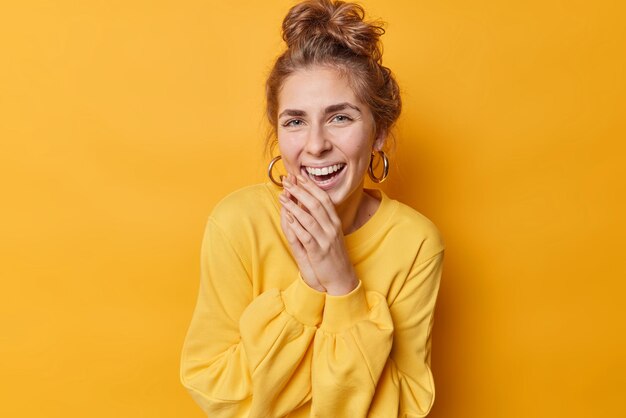 Fröhliche Europäerin hält die Hände in der Nähe des Mundes zusammen und lacht, als sie hört, wie etwas Lustiges in Freizeitkleidung gekleidet lächelt, isoliert über gelbem Hintergrund. Konzept der positiven Gefühle.